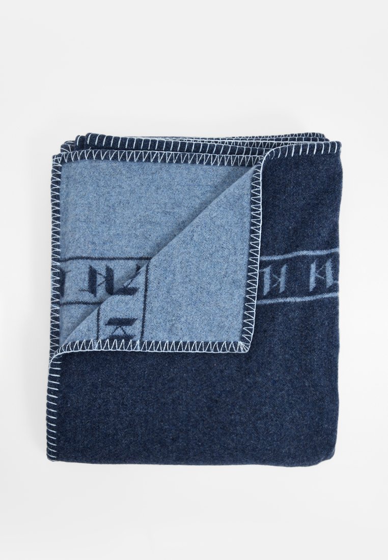 Couverture en laine avec un design bicolore en bleu foncé et bleu clair, avec des coutures visibles et un motif de logo le long du bord.
