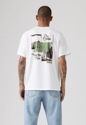 Homme avec des tatouages sur les bras, portant un t-shirt blanc avec un motif nature vert et noir et un jean bleu clair, tourné dos à la caméra.