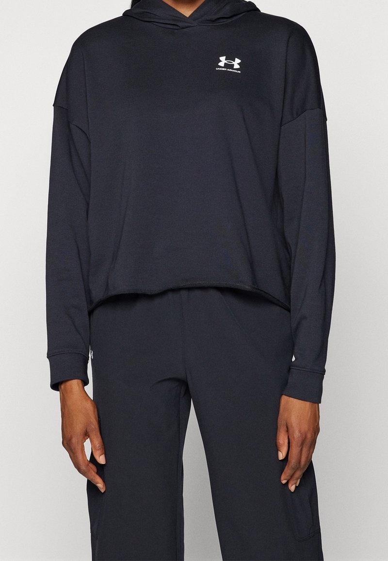 Personne portant un sweat à capuche noir court à manches longues et un pantalon noir assorti, avec un petit logo blanc Under Armour sur la poitrine.