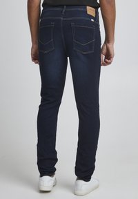 Jeans de hombre en azul oscuro hechos de denim, con bolsillos traseros con costura amarilla y una etiqueta de cuero marrón en la cintura. Diseño de corte ajustado.