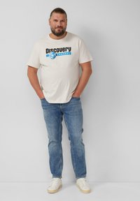 Cremfarbenes T-Shirt mit dem "Discovery Channel"-Logo in Schwarz und Blau. Kombiniert mit hellblauen Jeans und weißen Sneakern. Lässige Passform.