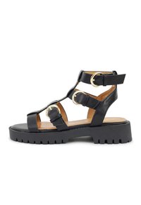 Schwarze Plateausandalen mit einer strukturierten Oberfläche, die über drei Riemenabschnitte mit goldenen, kreisförmigen Beschlägen und einer klobigen, geriffelten Sohle verfügen.