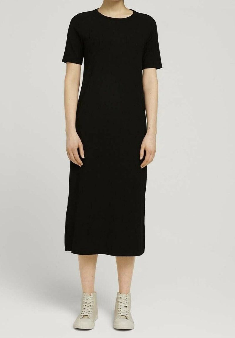 Personne portant une robe midi noire unie à manches courtes, associée à des baskets lacées de couleur claire, debout devant un fond neutre.