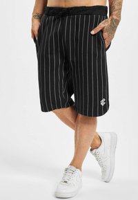 Pantaloni scurți negri cu dungi, cu talie elastică și șnur. Au buzunare laterale și un mic logo alb aproape de margine. Purtați cu adidași albi.