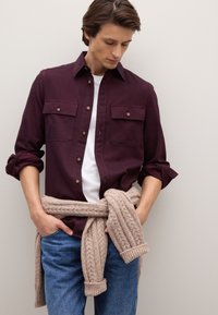 Bordeauxrotes Hemd mit Knöpfen und zwei Brusttaschen, kombiniert mit einem weißen T-Shirt. Beiger Strickpullover mit Zopfmuster um die Taille gebunden, blaue Jeans.