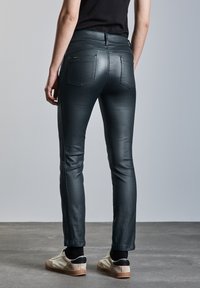 Persona vistiendo pantalones ajustados negros y zapatillas beige con calcetines negros, de pie sobre un suelo de concreto contra una pared gris clara.