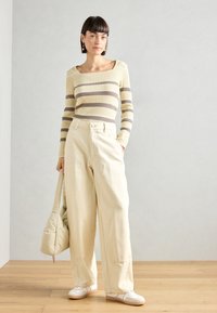 Maglietta a maniche lunghe, a righe gialle e marroni, con scollatura quadrata; pantaloni beige a gamba larga; sneakers bianche; borsa beige.
