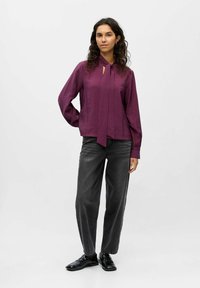 Lila Bluse mit einem Bindedetail, strukturiertem Stoff und langen Ärmeln, kombiniert mit locker sitzenden, dunkelgrauen Hosen und schwarzen Schuhen.