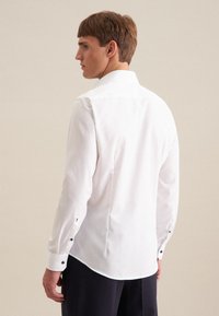 Camicia bianca con bottoni, maniche lunghe, vestibilità sartoriale e bottoni scuri a contrasto ai polsini; tessuto liscio, colletto classico, orlo curvo.