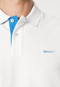 Weißes Poloshirt mit blauem Kragen, ausgestattet mit einer Knopfleiste mit zwei Knöpfen und einem kleinen blauen gestickten Logo auf der linken Brust.