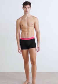Boxer-strings pour hommes noirs avec une taille rose comportant du texte en logo. Le tissu semble lisse et extensible, ajusté au corps.
