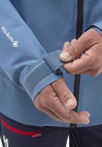 Blauer wasserdichter Jacke mit einem Velcro-Manschettenriemen, strukturiertem Design und schwarzem Reißverschlussdetail. Hände passen die Manschette an, was die Funktionalität betont.