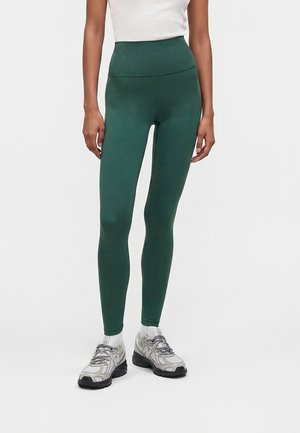 Personne portant des leggings taille haute verts foncés, un top blanc sans manches, des chaussettes blanches et des chaussures de sport grises et blanches, debout sur un fond uni.