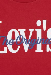 Κόκκινο βαμβακερό μπλουζάκι με έντονα λευκά και μπλε γράμματα που αναγράφουν χαρακτηριστικά "Levi's" και "The Original". Ομαλή υφή.