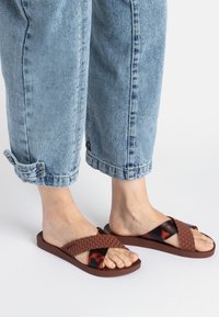 Braune gewebte Rutschsandalen mit einem glänzenden Schildpattmuster auf dem Riemen, kombiniert mit hochgekrempelten, hellblauen Jeans.