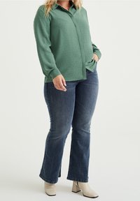 Grön skjorta med knappar och texturerat mönster, parat med utsvängda blå jeans och beige ankelstövlar. Skjortan har krage och långa ärmar.