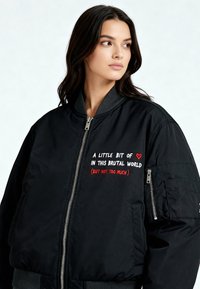 Zwarte bomberjack met een ritssluiting aan de voorkant, geribde kraag en geborduurde tekst in wit en rood. Voorzien van zijzakken en een gestructureerde afwerking.