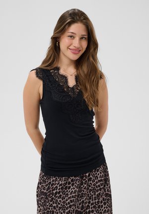 Donna sorridente con lunghi capelli castani che indossa un top smanicato in pizzo nero e una gonna con stampa leopardata, in piedi davanti a uno sfondo semplice.