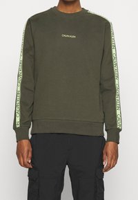 Sudadera verde oliva con puños y dobladillo acanalados. Presenta un logo amarillo de "Calvin Klein" a lo largo de los hombros y las mangas.
