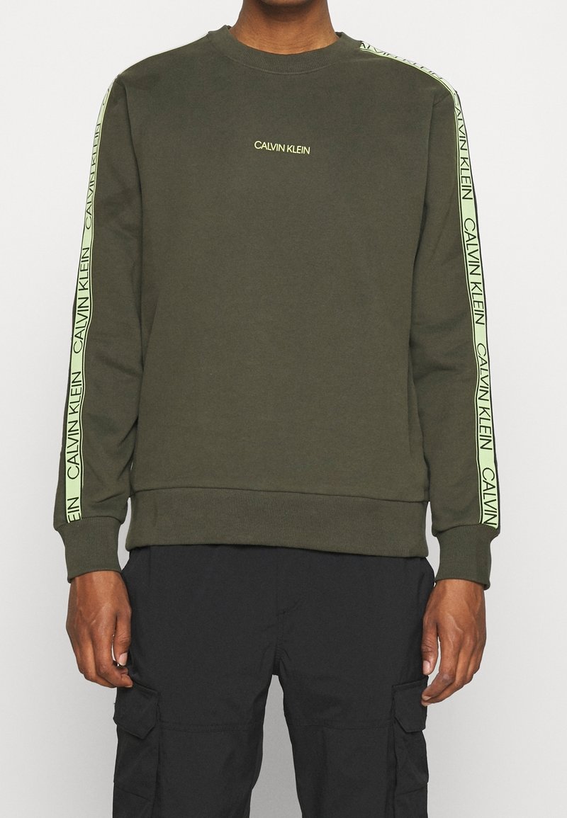 Sudadera verde oliva con puños y dobladillo acanalados. Presenta un logo amarillo de "Calvin Klein" a lo largo de los hombros y las mangas.
