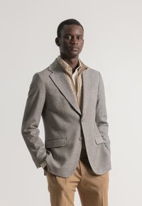 Blazer gris a cuadros con solapas tipo notches y dos bolsillos frontales, superpuesto a una chaqueta beige con cremallera. Pantalones marrones completan el conjunto.