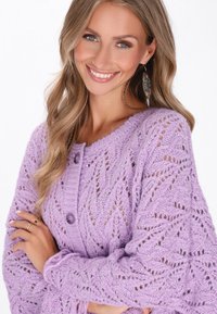 Ljuslila stickad kardiagan med ett zigzag-mönster, två stora knappar och texturerade, öppna stickmönster. Mjuk och mysig utseende.