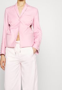 Roze blazer met zijuitsparingen, lange mouwen en een voorclosure met knopen. Gecombineerd met lichtroze broeken. Glad materiaal met minimale details.