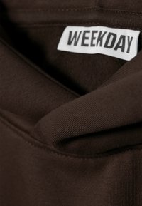 Tela de algodón marrón con una textura suave, que presenta una etiqueta visible que dice "WEEKDAY". La tela tiene un diseño simple y sin adornos.