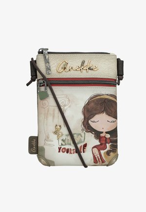 Bolso bandolera pequeño de color crema con una niña de dibujos animados con vestido rojo, logo "Anekke", texto "Conócete a ti mismo" y un charm de gato en la cremallera frontal.