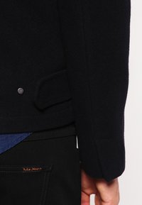 Marineblaue Wollmischjacke mit schmaler Passform, mit einer Seitentasche und einem silbernen Druckknopfdetail, kombiniert mit dunklen Jeans.