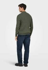 Pull en maille torsadée vert avec un col rond, des manches longues et des poignets côtelés, associé à un jean bleu foncé et des chaussures noires.
