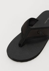 Sandal cu vârf flip-flop negru, cu talpă din cauciuc texturat. Are o curea din țesătură în formă triangulară cu logo-ul CK negru. Design simplu și funcțional.