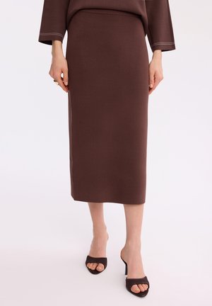 Kobieta nosząca brązową, dopasowaną, średniej długości spódnicę oraz pasujący top z długim rękawem, zestawione z ciemnobrązowymi, otwartymi sandałami na wysokim obcasie.