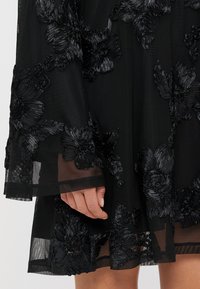 Melnas ziedu izšūšanas kleita ar caurspīdīgu audumu. Piedāvā sarežģītus melnus ziedu rakstus uz piedurknēm un apakšmalas, uzsverot faktūru un detaļas.