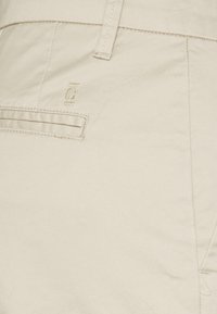 Pantaloni in cotone beige con una finitura texturizzata, dotati di tasca posteriore e logo ricamato in vita, che mostrano un design semplice e classico.