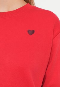 Felpa rossa con scollatura tonda e polsini a coste. Presenta un piccolo cuore scuro ricamato sulla parte sinistra del petto, realizzata in tessuto morbido.