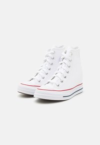 White canvas high-top sneakers with rounded toe, metallic eyelets, flat laces, red and blue accents on the sole. Textured sole for grip.