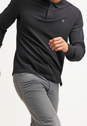 Zwart poloshirt met lange mouwen, een kraag en twee knopen, met een klein logo op de borst. Gedragen met grijze spijkerbroek, in een actieve pose.
