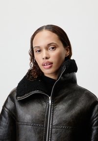 Giubbotto di pelle nera con una finitura testurizzata, caratterizzato da un colletto in peluche nero e una zip argentata. Il design è oversize e strutturato.