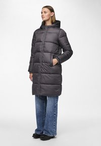 Lang grå dunjakke med hette, med glidende frontlås, sidelommer og quiltet design. Brukt med vide jeans og svarte sko.