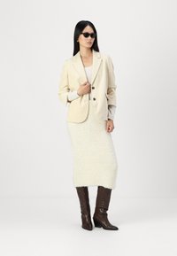 Beige korduroyjakk med to knapper, lagd over en lys genser og et kremfarget strikket skjørt som går til kneet. Brune lærstøvler som går til knéet.