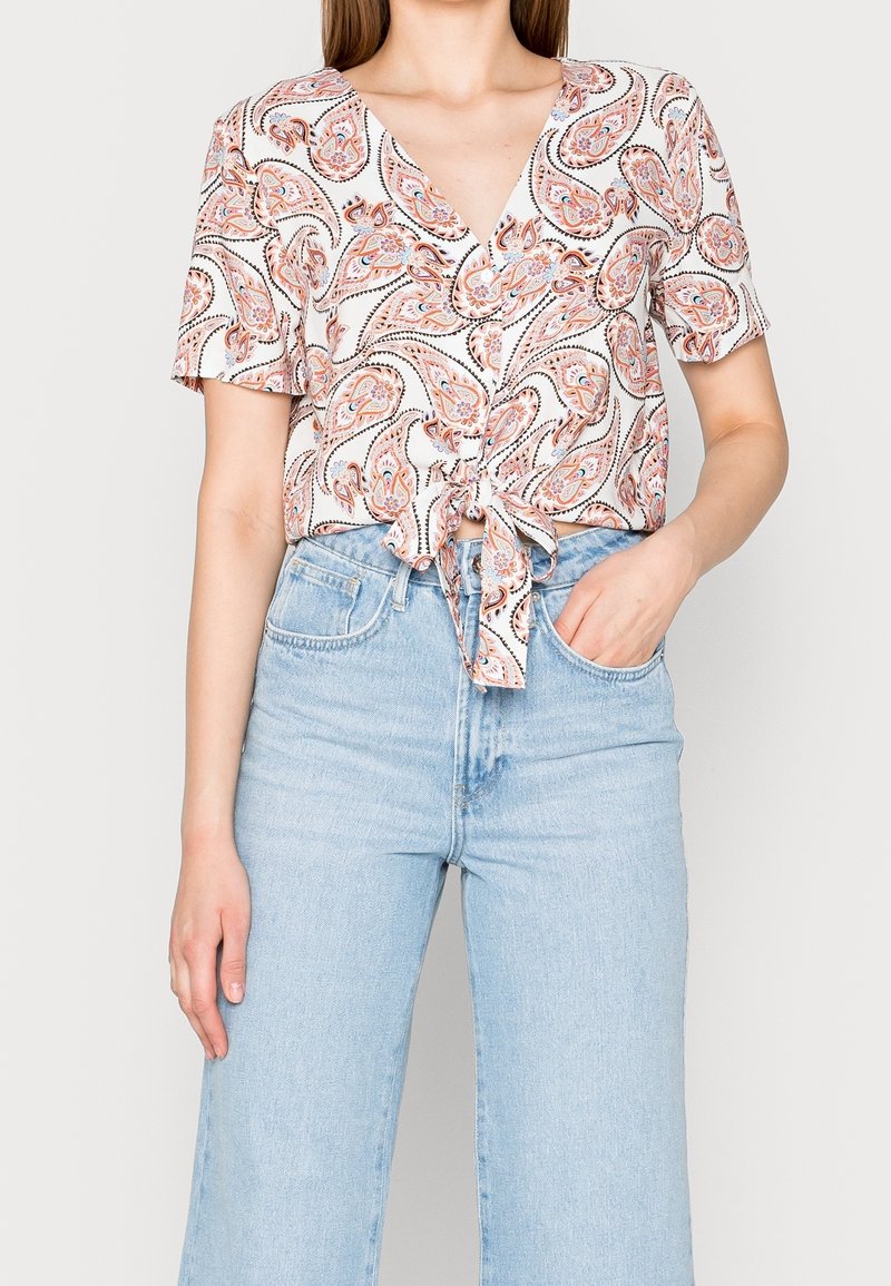 Donna che indossa un top a maniche corte con motivo paisley legato in vita, abbinato a jeans a vita alta di colore blu chiaro, con una mano in tasca.