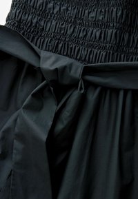 Musta mekko, jossa on smokkattu bodysi, solmittu vyötäröyksityiskohta ja liehuva helma. Kangas näyttää kevyeltä ja sileäpintaiselta.