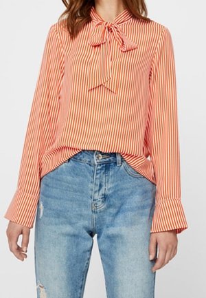 Frau trägt eine orange-weiß vertikal gestreifte Bluse mit Schleifenkragen und blaue, hoch taillierte, verwaschene Jeans.