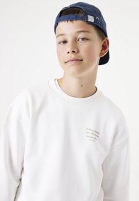 Sudadera blanca con cuello redondo y tejido texturizado. Presenta texto bordado en color beige en la parte delantera. Combinada con una gorra de béisbol azul.