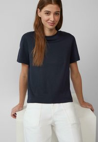 Granatowa koszulka z krótkim rękawem z okrągłym dekoltem, wykonana z miękkiego materiału, w połączeniu z białymi spodniami o wysokim stanie, stojąca na neutralnym tle.