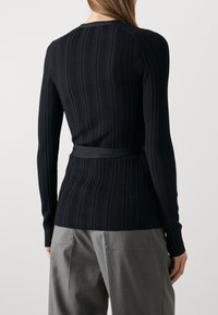 Svart ribbad långärmad topp med en figursydd silhuett och ett knytbälte i midjan. Manschetterna är något insvängda.