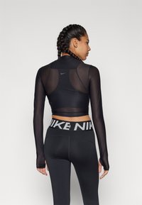 Femeie cu păr împletit, îmbrăcată cu un top scurt din plasă neagră cu mânecă lungă și ciorapi Nike negri cu un logo îndrăzneț pe talie, stând pe un fundal alb.
