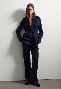 Blazer doppiopetto blu navy con reversing a punta, abbinato a pantaloni su misura dello stesso colore. Tessuto liscio con un leggero lucido e una vestibilità strutturata.