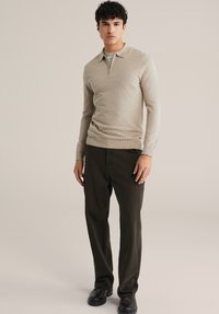 Bézs színű, kötött pulóver cipzáras gallérral, sötétzöld nadrággal és fekete cipővel viselve. A szövet finom textúrájú és alakra szabott kialakítású.